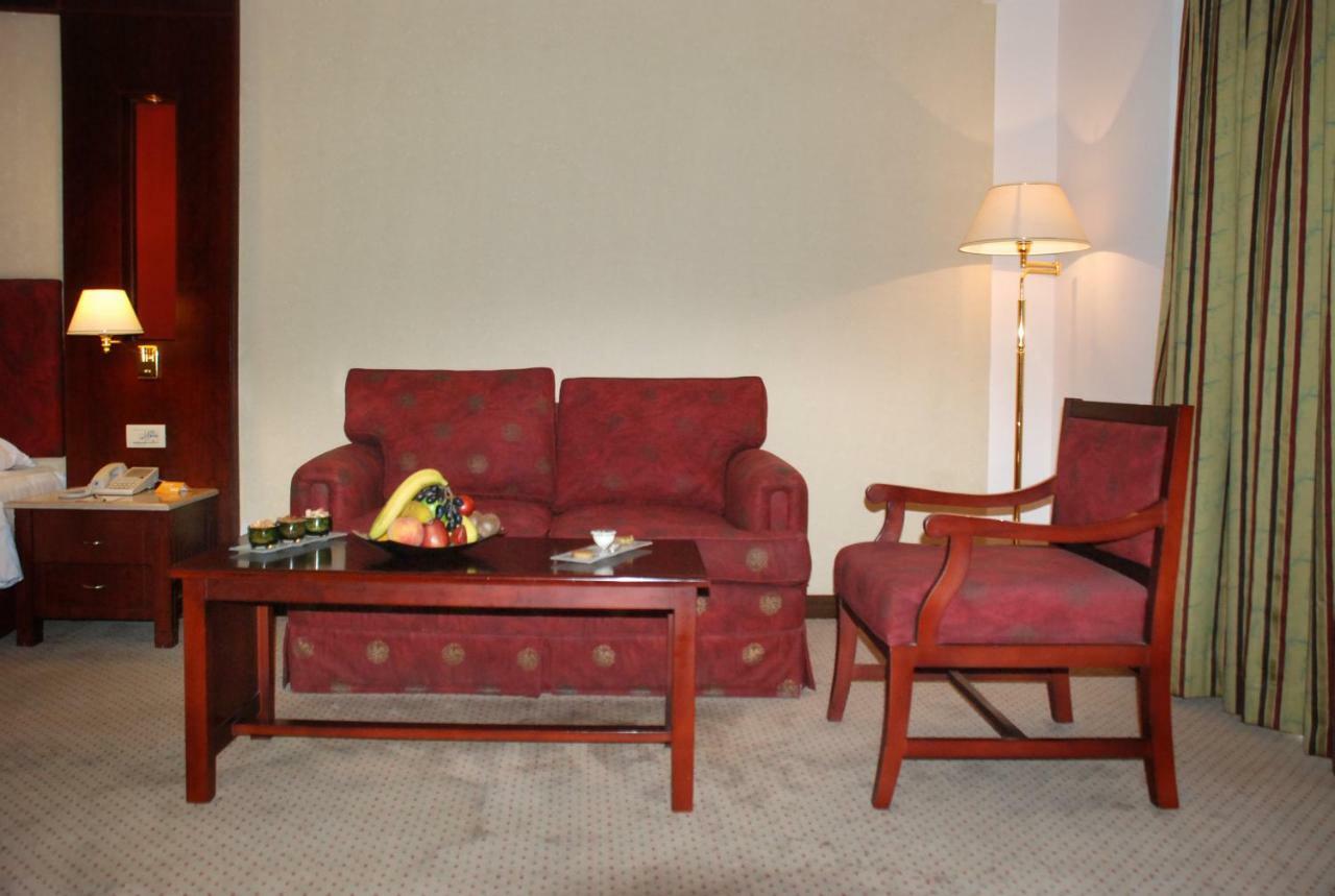 โรงแรมซาเฟอไคโร Giza ภายนอก รูปภาพ The photo shows a cozy seating area in a hotel room. There is a red patterned sofa and a matching armchair placed around a wooden coffee table. On the table, there are some snacks or fruits displayed. A floor lamp is positioned beside the sofa, and t
