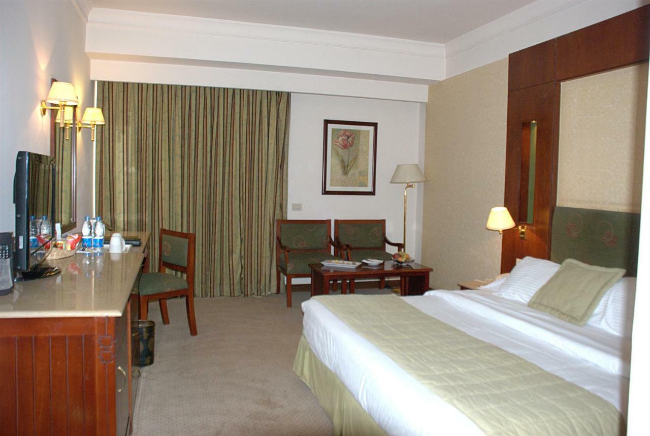 โรงแรมซาเฟอไคโร Giza ห้อง รูปภาพ The photo shows a hotel room. It features a bed with white linens and a green throw. On one side of the bed, there's a small bedside lamp. Opposite the bed is a desk with a mirror above it, alongside a couple of chairs and a small table. The room has